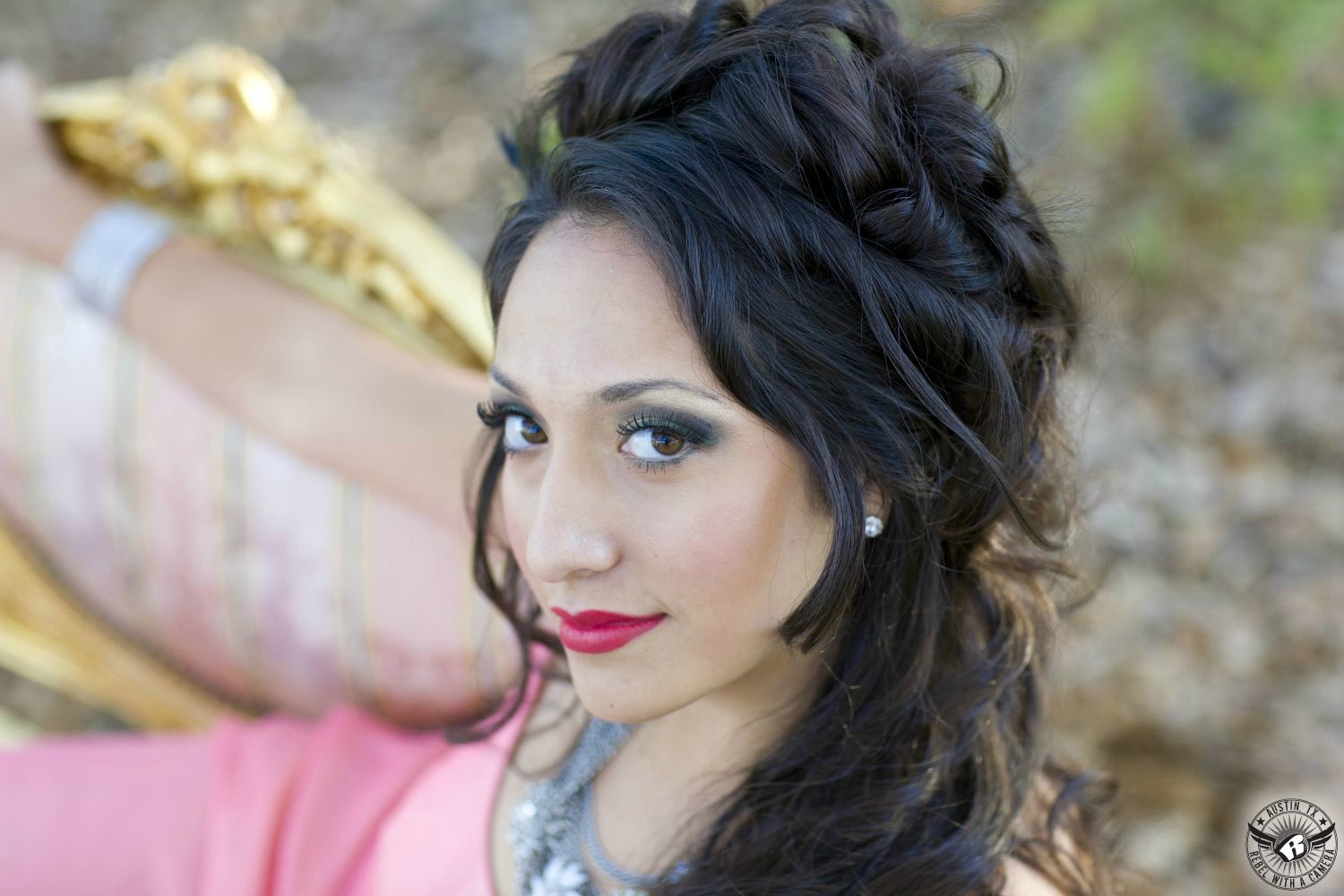 ethnic brunette model with curly hair and brown eyes with bright lipstick with a pink dress and lots of bringy necklaces sitting on out of focus vintage gold couch outside in the fall  leaves and soft light